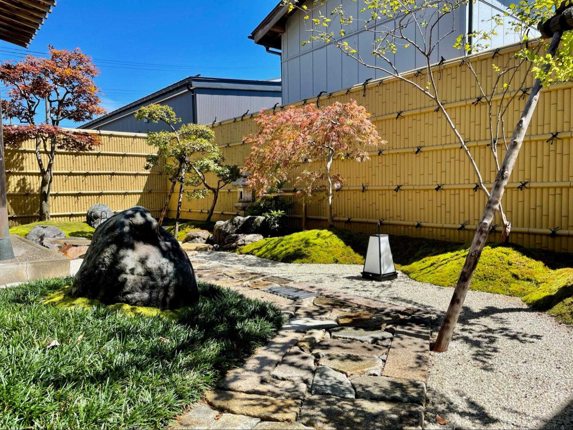 施工後の和風庭園。竹垣沿いに苔の築山と大きな庭石、赤いモミジなどの庭木を配した昼の様子